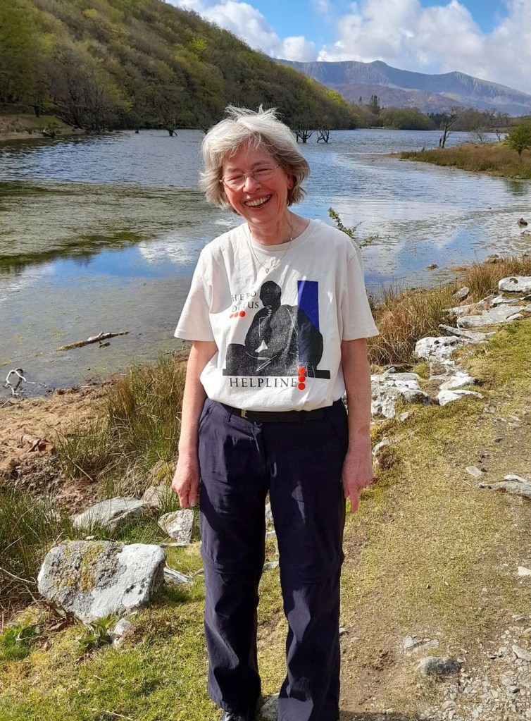 Doris Post, an older woman with short hair wearing a Herodotus Helpline t-shit, smiles broadly at the camera, backed by a gorgeous natural scene including a winding river, several mountains, and a blue sky with puffy clouds.